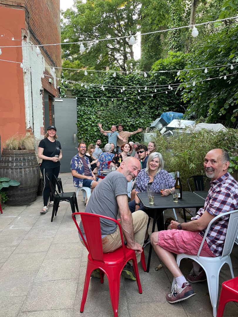 The Barden: Smiling member customers seated in the yard garden bar, the Barden, with greenery and festoon lighting