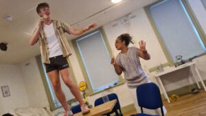 Sam stands on the table whilst Kananu is on the ground whilst they are singing into wooden spoons