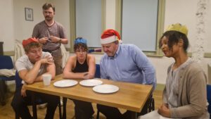 The entire cast sit around a table in christmas hats 