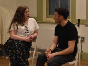 Male and female actor sit facing each other, in conversation