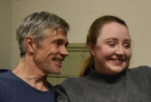 Close-up of male actor sitting looking sideways at female actor, both laughing