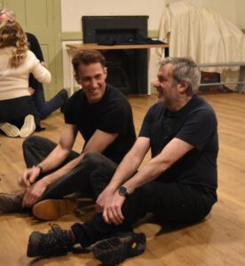 Two male actors sit on the floor laughing at a shared joke, with other actors in background