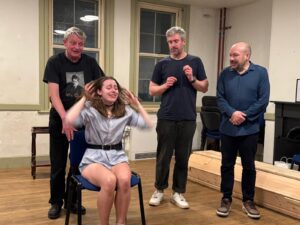 A woman sits on a chair in distress, surrounded by three men standing