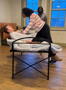 A woman stands over a man shocked and supine on a bed with her hands on his chest, while another man looks on