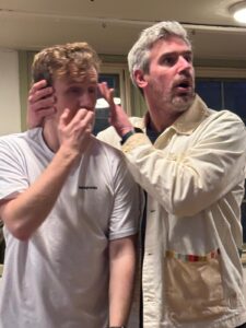 Two men standing one looks shocked and blocks the ears of the other