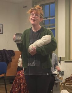 Smiling female actor with a babe in arms holds a mug of beer