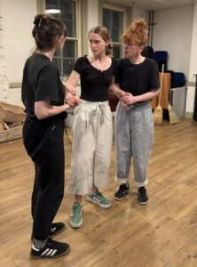 Three female actors stand holding hands in a tense, emotional scenes