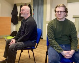 Two male actors sit facing away from each other, hands in laps