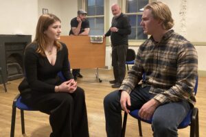 Male and female actor sit talking together, while two male actors stand looking on in the background