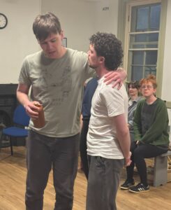 Two male actors stand in conversation, one with his arm around the other conspiratorially