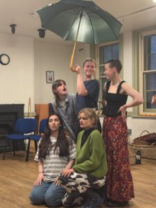 Five female actors stand and crouch beneath a large green umbrella