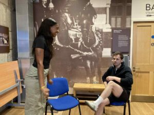 A woman stands looking and smiling at a man sitting in a relaxed, slightly arrogant pose