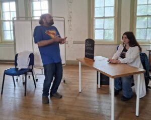 A man standing with hands outstretched towards a woman in a white coat sitting at a desk, looking askance at him