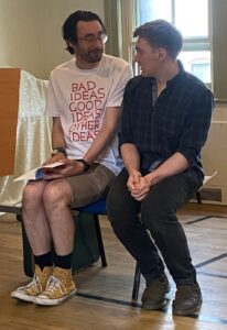Two male actors sit closely together, chatting happily