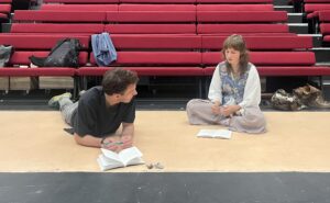 Male actor lies on floor propped up on elbows talking to female actor sitting cross-legged beside him