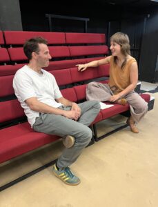 A male and female actor sit facing each other on a bench seat., laughing