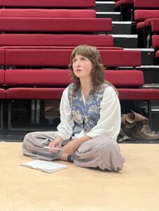 A female actor sits cross-legged on the floor, looking upwards