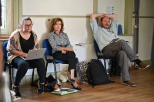 Two women and a man sit on the periphery of the rehearsal room, watching the action beyond