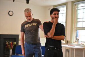 A man stands with his hand to his mouth in distress; another man stands behind him, putting a comforting hand on his shoulder