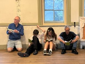 Two male and a female actor sit in a row reading their scripts beneath a window. A third male actor kneels alongside them, looking right