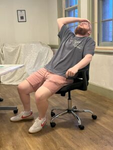 A male actor sits back in a swivel chair clutching his head as though in despair