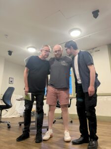 Three male actors stand close together in a row; the one in the centre has his arms around the shoulders of the other