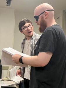 Two actors stand in conversation, one wearing an eye patch, the other with a surprised expression