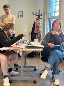 A male actor in a bad wig and an eye patch sits across a desk from and points at a female actor sitting in a defensive pose. A second male actor looks on behind them