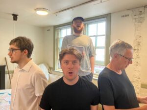Three actors stand in a line singing, with a fourth behind them at a height