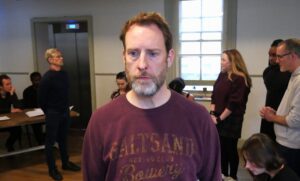 Male actor with worried expression stands in foreground with others behind him talking