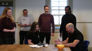 A male and female actor sit at a table in serious conversation with three men and a woman looking on behind them with concern