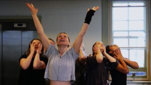 Five young female actors stand in ecstasy, with one at the front with her arms joyously aloft