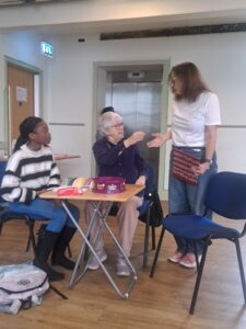 An older actor sitting at a table gives something to one standing next to her, while a young actor looks on