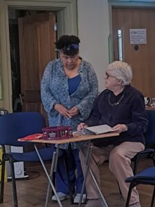 An older actor sits at a table, while her partner stands at her shoulder