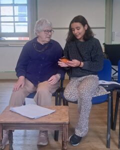 A young actor is showing an older one something on a mobile phone, both seated and relaxed