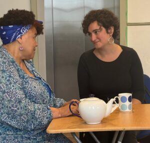 Two actors sit close together at a table with teapot and cups