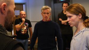 A young female actor is questioned by two men, while others look on in the background