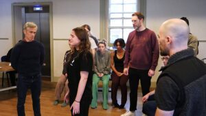 A distraught young woman is questioned by an older man,, while several actors look on around them 