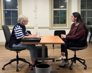Two female actors look intently at each other, sitting across a desk from each other as though in an office 