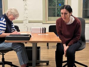 A female actor sits on an office chair in some distress, while another sits across from her making notes and not looking up