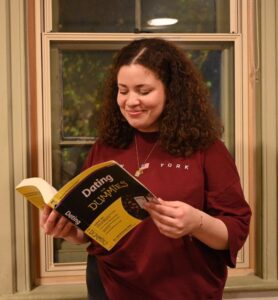 A young woman stand smiling as she reads a book on Dating