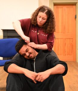A young woman sits in a chair combing the hair of a young man with a pained expression who sits at her feet