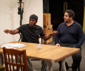 Two men sit a table, one points out to the side while explaining something to the other