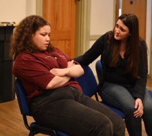Two female actors sit in deep conversation, one with her arms folded defensively