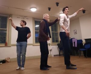 Three male actors stand as though holding lanterns, looking around them