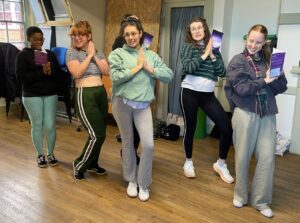 Five female actors stand posing happily looking to the front of the room with hands in prayer