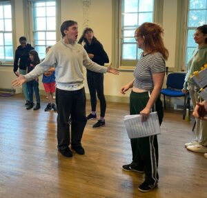 A male actor stands with arms outstretched while a mix of female and child actors look on