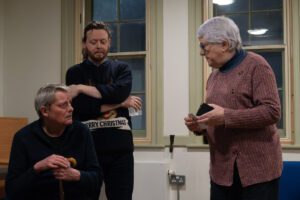 Female actor with notebook in hand stands talking to a seated male actor, with another man standing behind him, arms folded