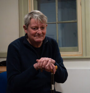 A male actor sits in front of a window, leaning on a walking stick, looking down with a sad expression