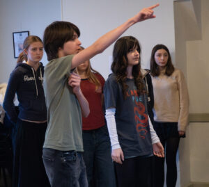 Six young actors standing in rehearsal, one pointing,the other looking in that same direction